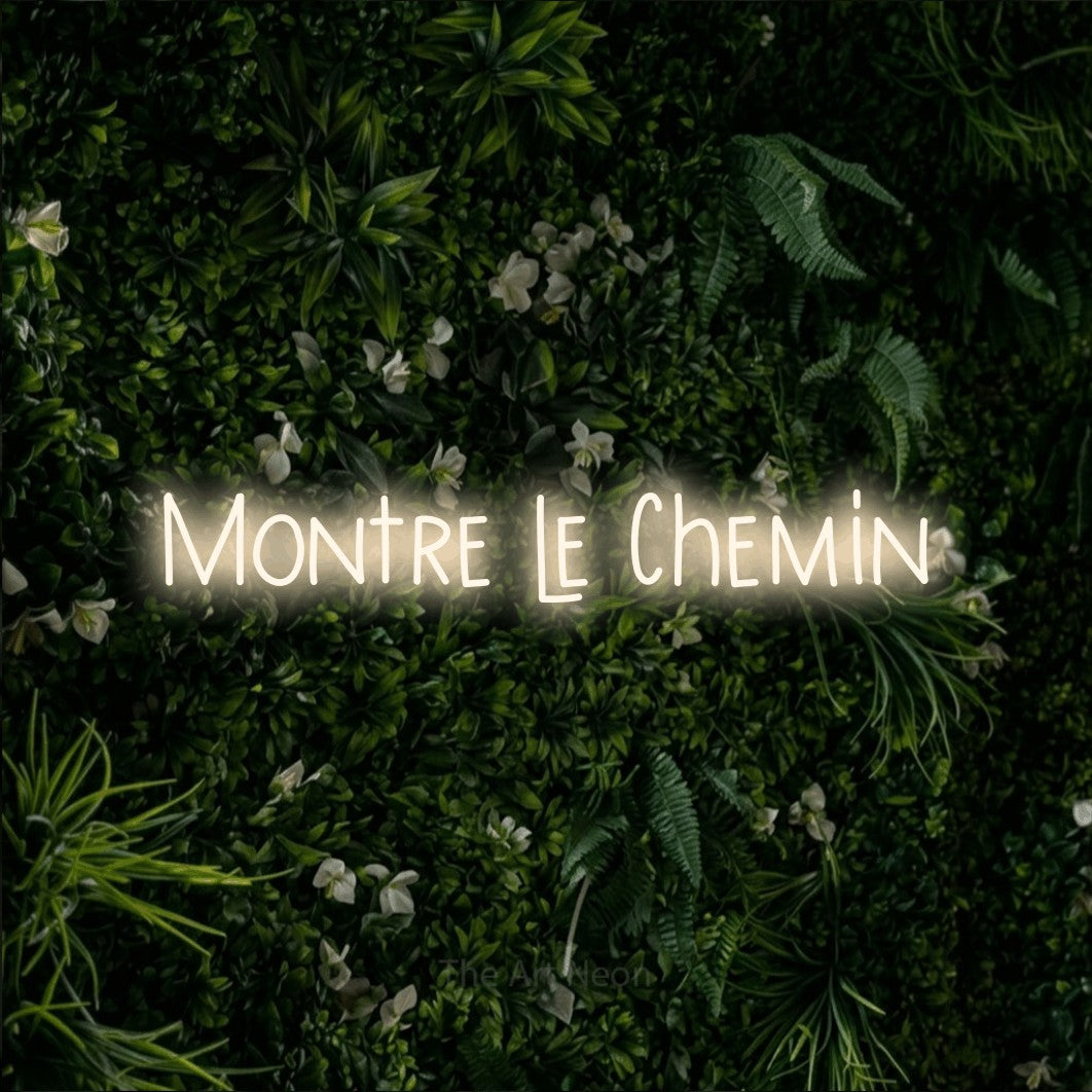 Aperçu d’une enseigne néon LED « Montre le chemin » en blanc chaud, en lettres étroites dessinées à la main, photographiée sur un mur végétal vert et dense, pour un bureau ou une entreprise.