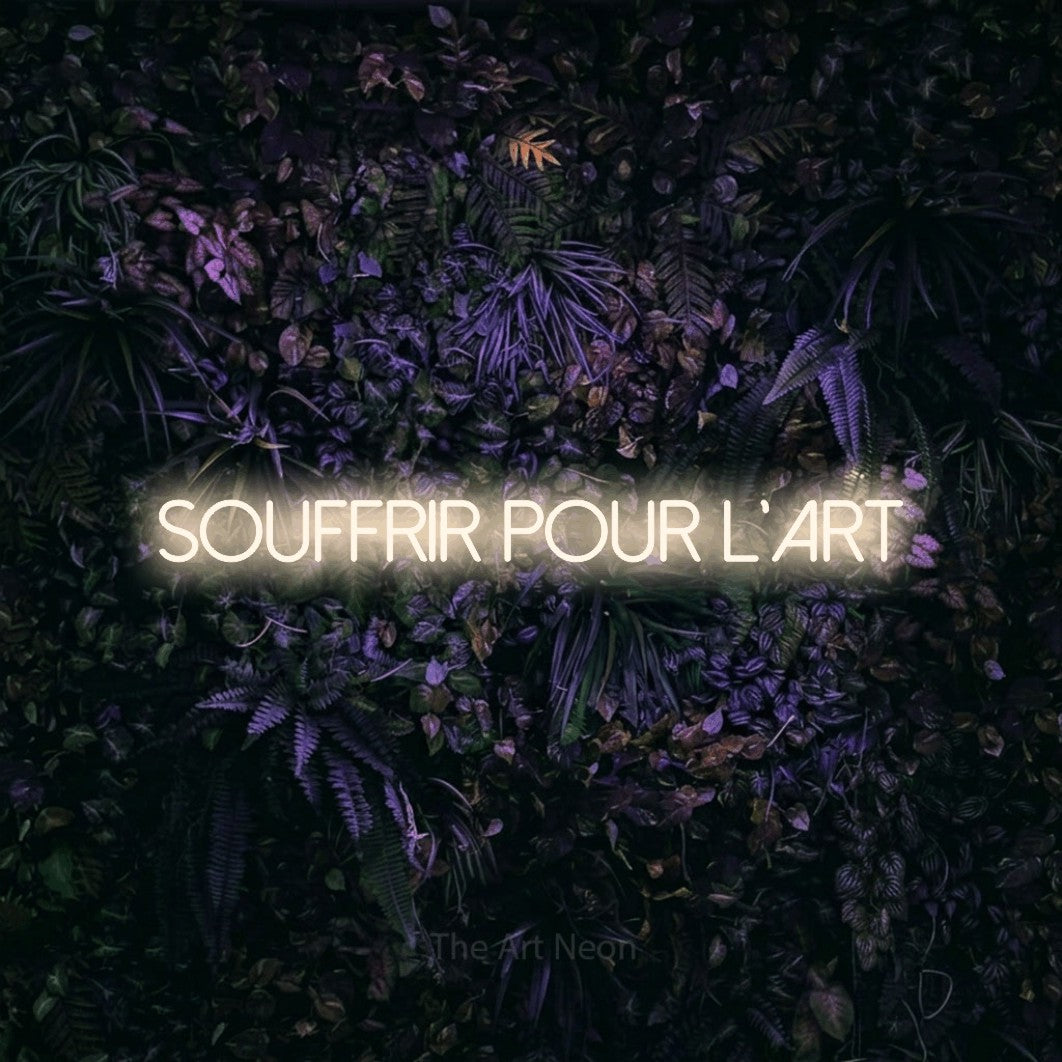 “Souffrir pour l'art” LED neon sign in warm white, with thin sans-serif lettering, shown against a dark wall with purple flowers.