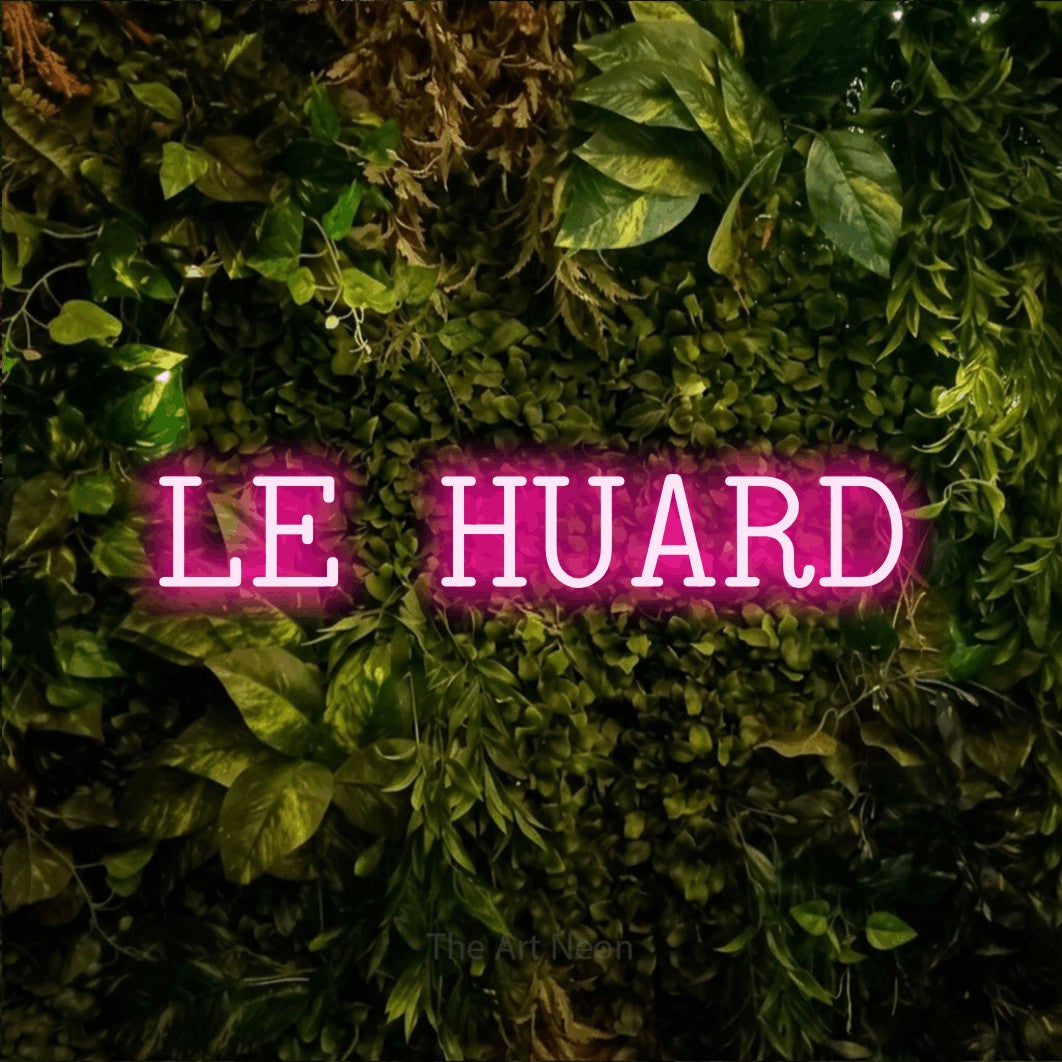 “Le huard” LED neon sign in hot pink, with typewriter-style lettering, shown against bright green and yellow foliage.