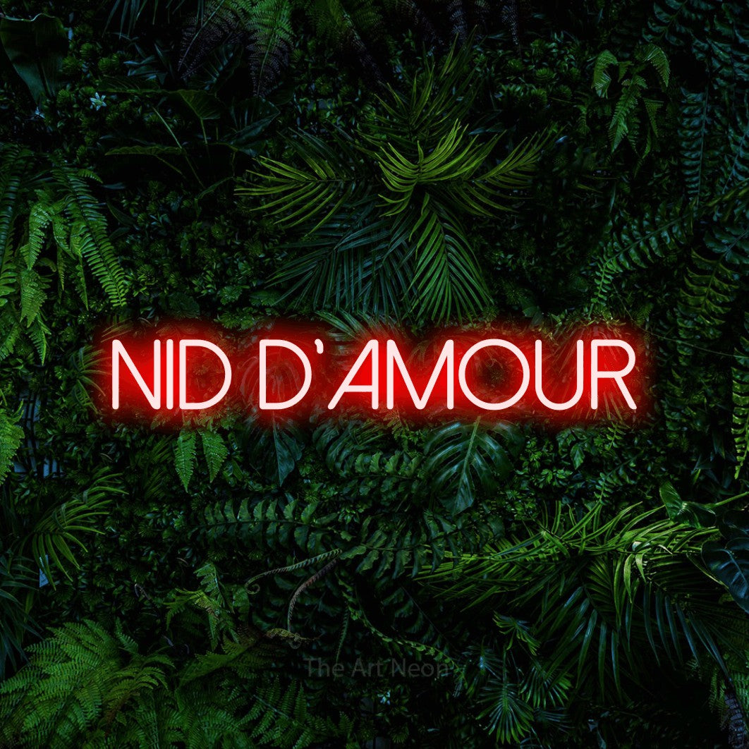 “Nid d'amour” LED neon sign in red, with thin sans-serif lettering, shown against dense tropical foliage.