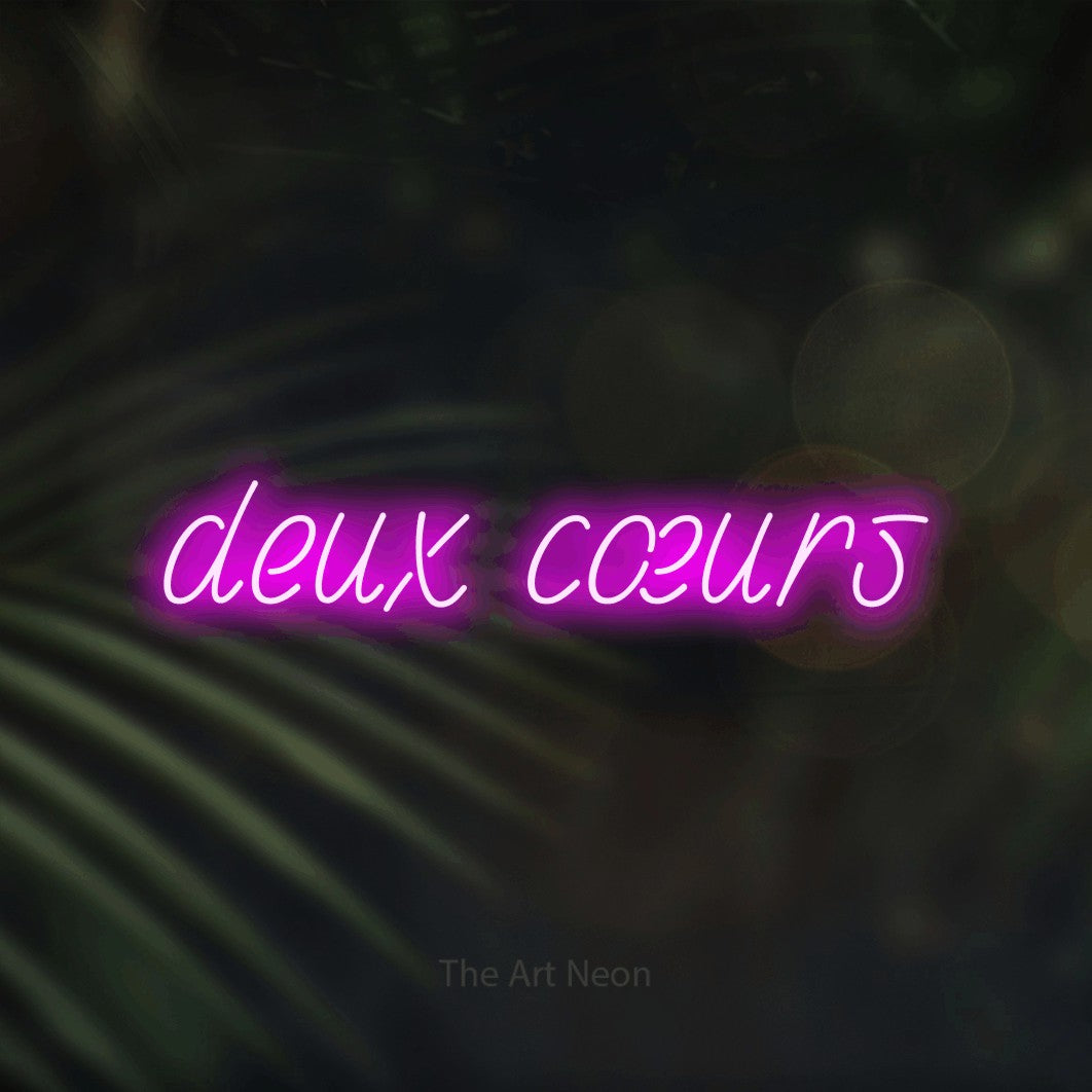 “Deux cœurs” LED neon sign in pink, with slanted handwritten lettering, shown against a blurred palm and bokeh-light backdrop.