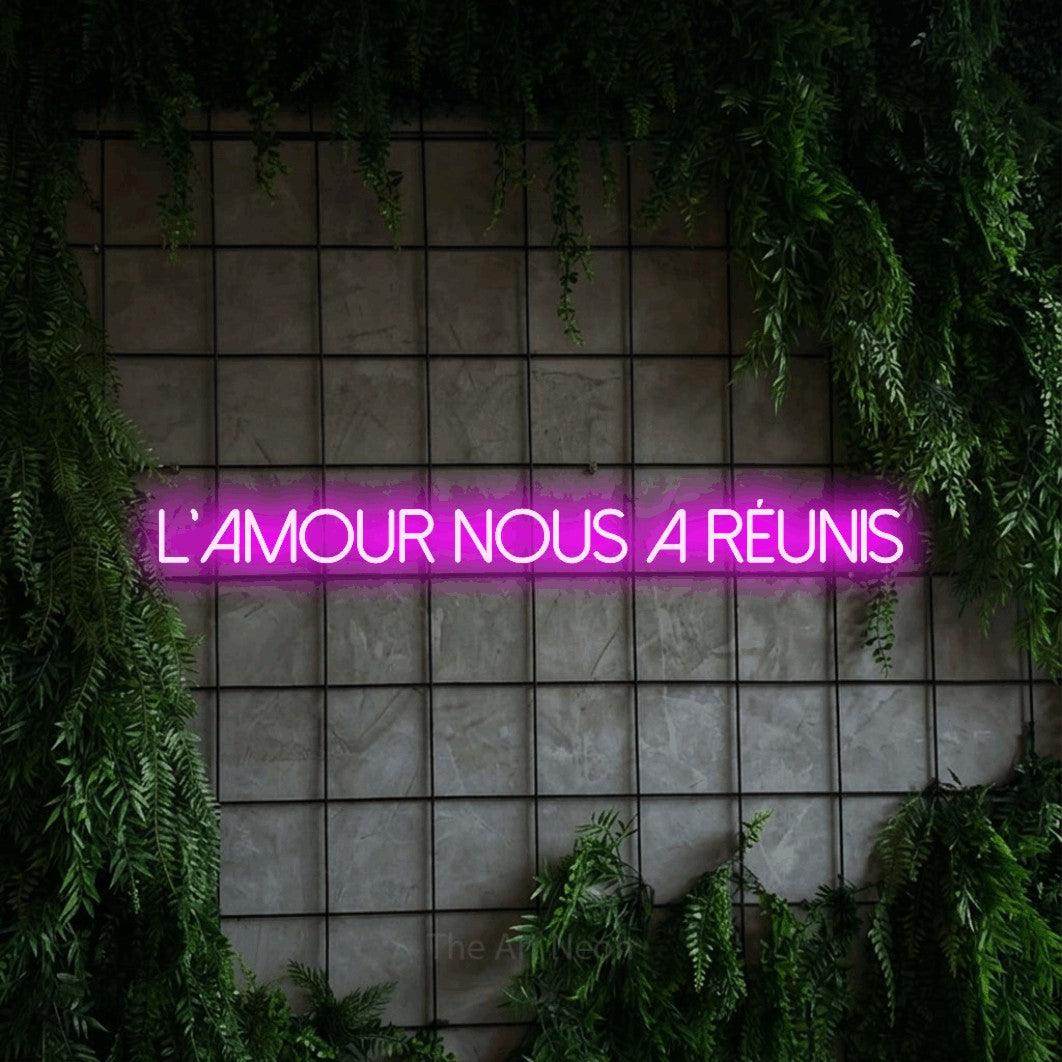 “L'amour nous a réunis” LED neon sign in pink, with thin sans-serif lettering, shown against an ivy-covered concrete grid.