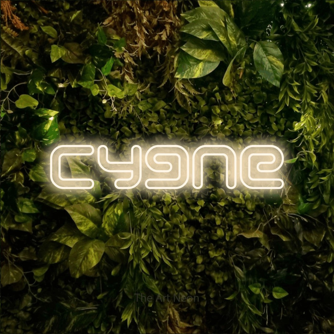 “Cygne” LED neon sign in warm white, with futuristic double-line lettering, shown against bright green and yellow foliage.