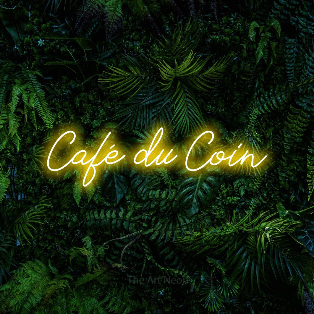 “Café du coin” LED neon sign in yellow, with cursive lettering, shown against dense tropical foliage.