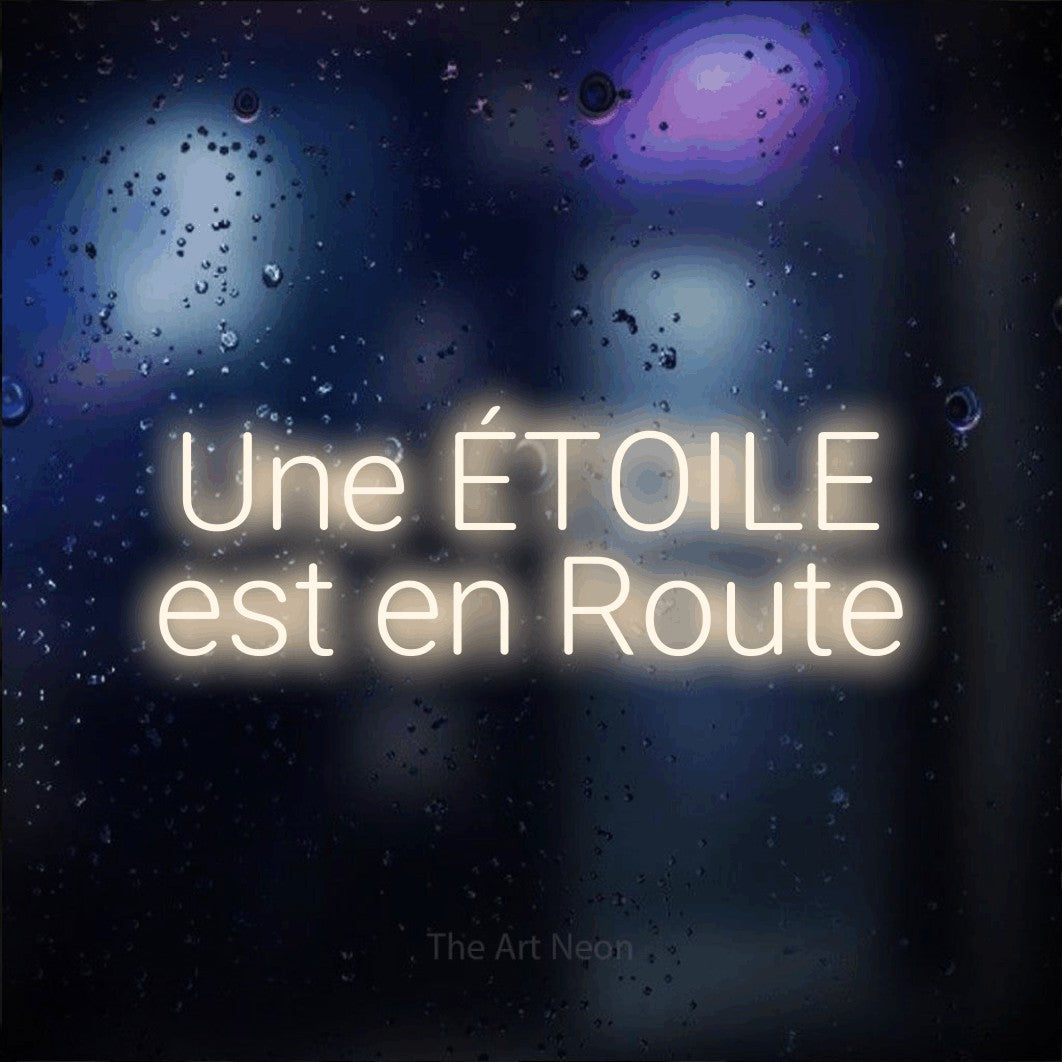 Néon LED « Une étoile est en route » sur deux lignes en blanc chaud, en lettres sans empattement arrondies, mis en scène sur une vitre aux reflets bleus et violets, pour une fête prénatale.