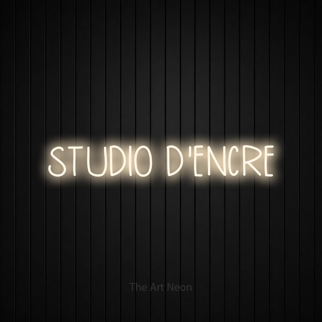 “Studio d'encre” LED neon sign in warm white, with narrow hand-drawn lettering, shown against black vertical wall slats.