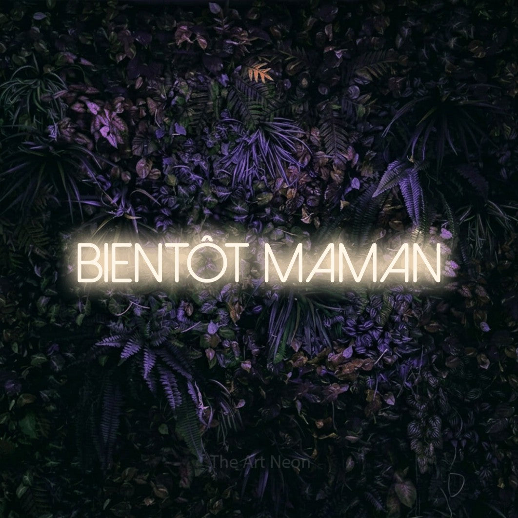 Néon LED « Bientôt maman » en blanc chaud, en lettres fines sans empattement, mis en scène sur un mur sombre aux fleurs violettes, pour une fête prénatale.
