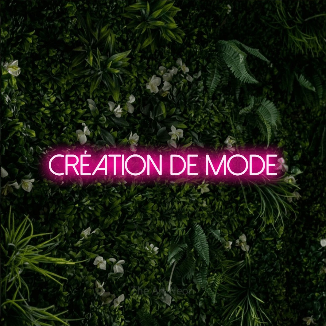 “Création de mode” LED neon sign in hot pink, with thin sans-serif lettering, shown against a dense green plant wall.