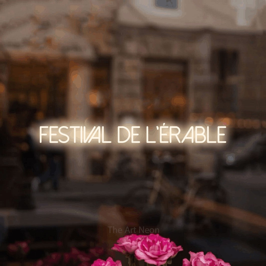“Festival de l'érable” LED neon sign in warm white, with thin sans-serif lettering, shown against a blurred café-bar interior.