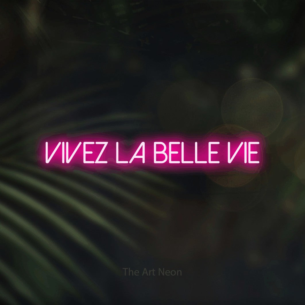 “Vivez la belle vie” LED neon sign in hot pink, with thin sans-serif lettering, shown against a blurred palm and bokeh-light backdrop.