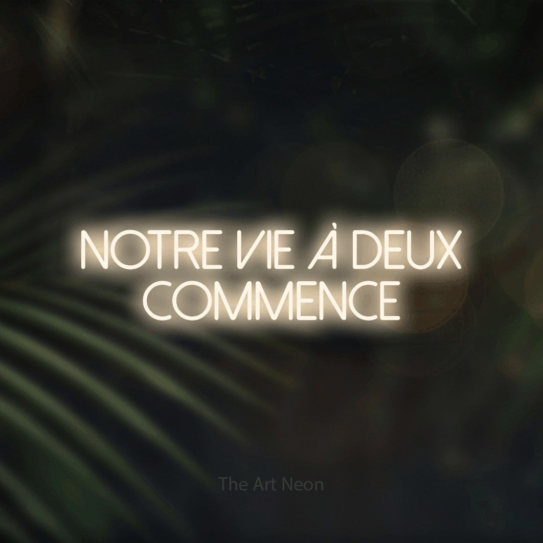 “Notre vie à deux commence” LED neon sign in warm white, with thin sans-serif lettering across 2 lines, shown against a blurred palm and bokeh-light backdrop.