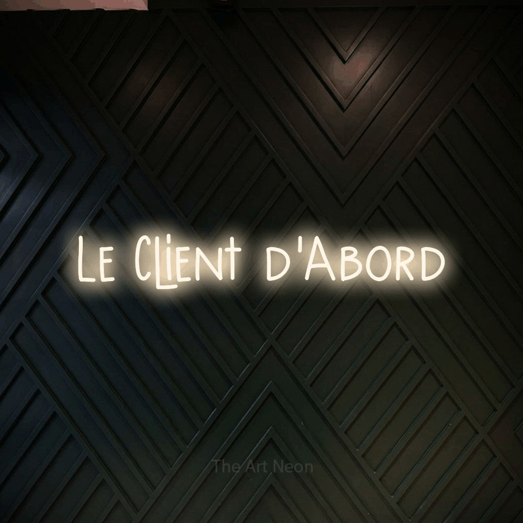 Lettrage en néon LED « Le client d'abord » en blanc chaud, en lettres étroites dessinées à la main, présenté sur un mur en bois noir à motif chevron, pour un bureau ou une entreprise.