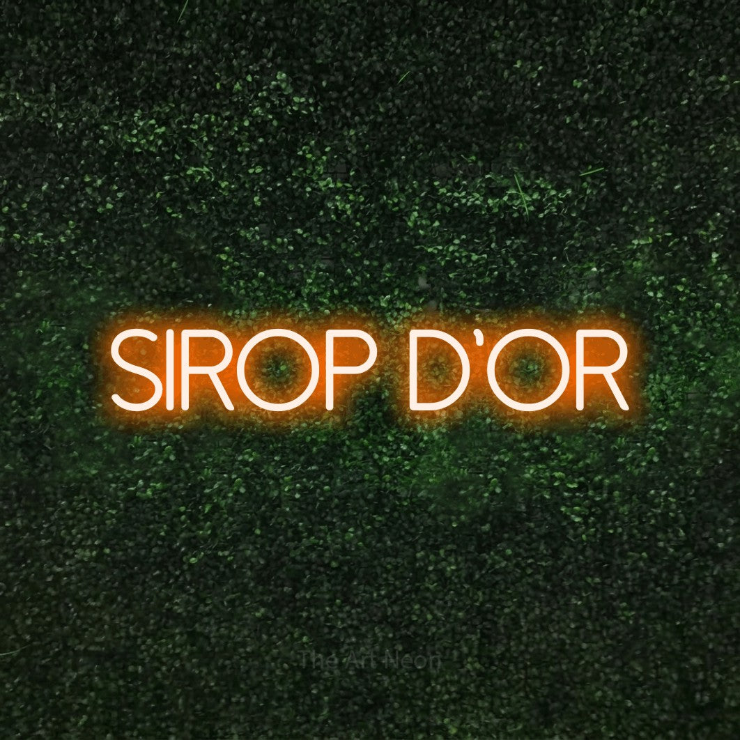 “Sirop d'or” LED neon sign in orange, with thin sans-serif lettering, shown against a textured green hedge.