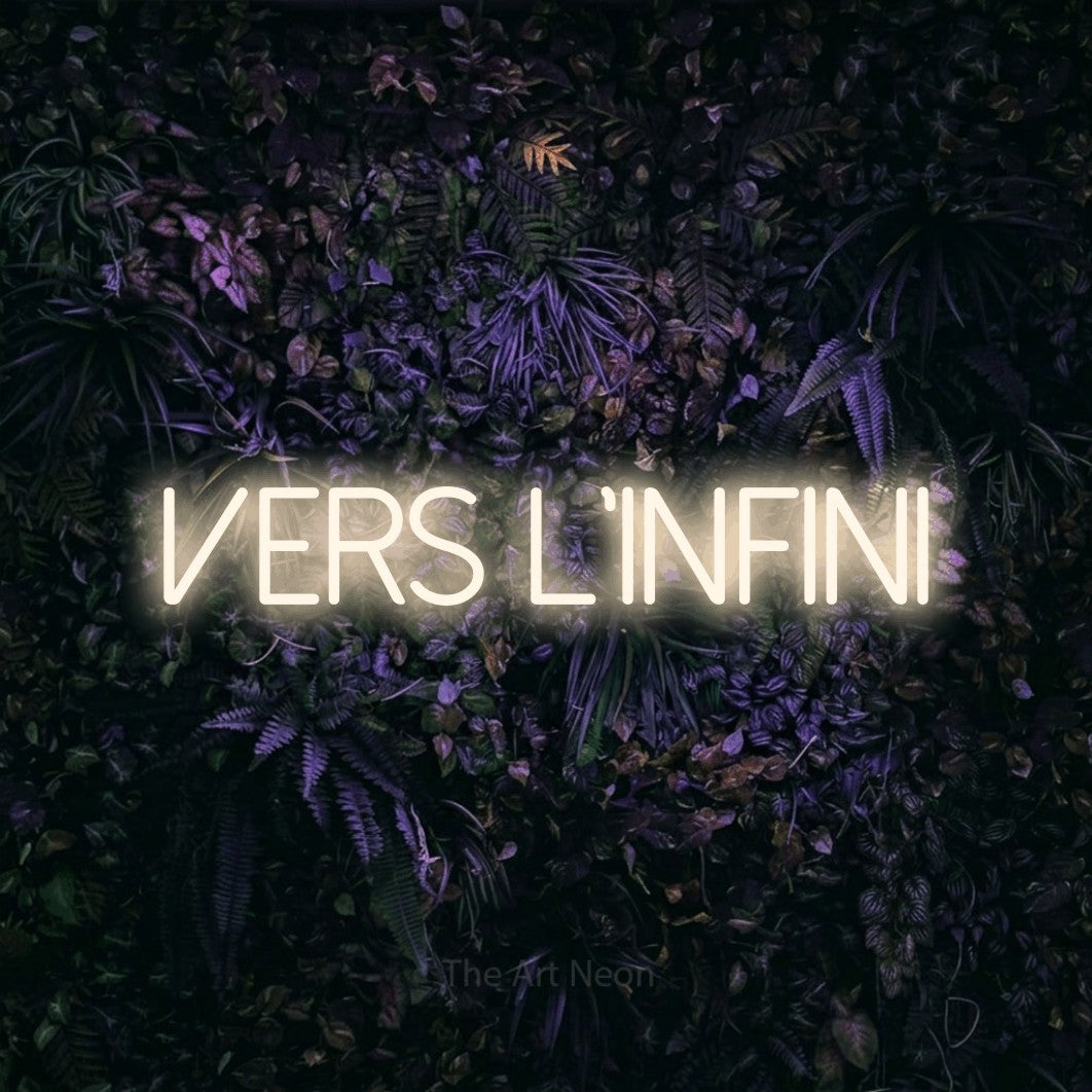 “Vers l'infini” LED neon sign in warm white, with thin sans-serif lettering, shown against a dark wall with purple flowers.