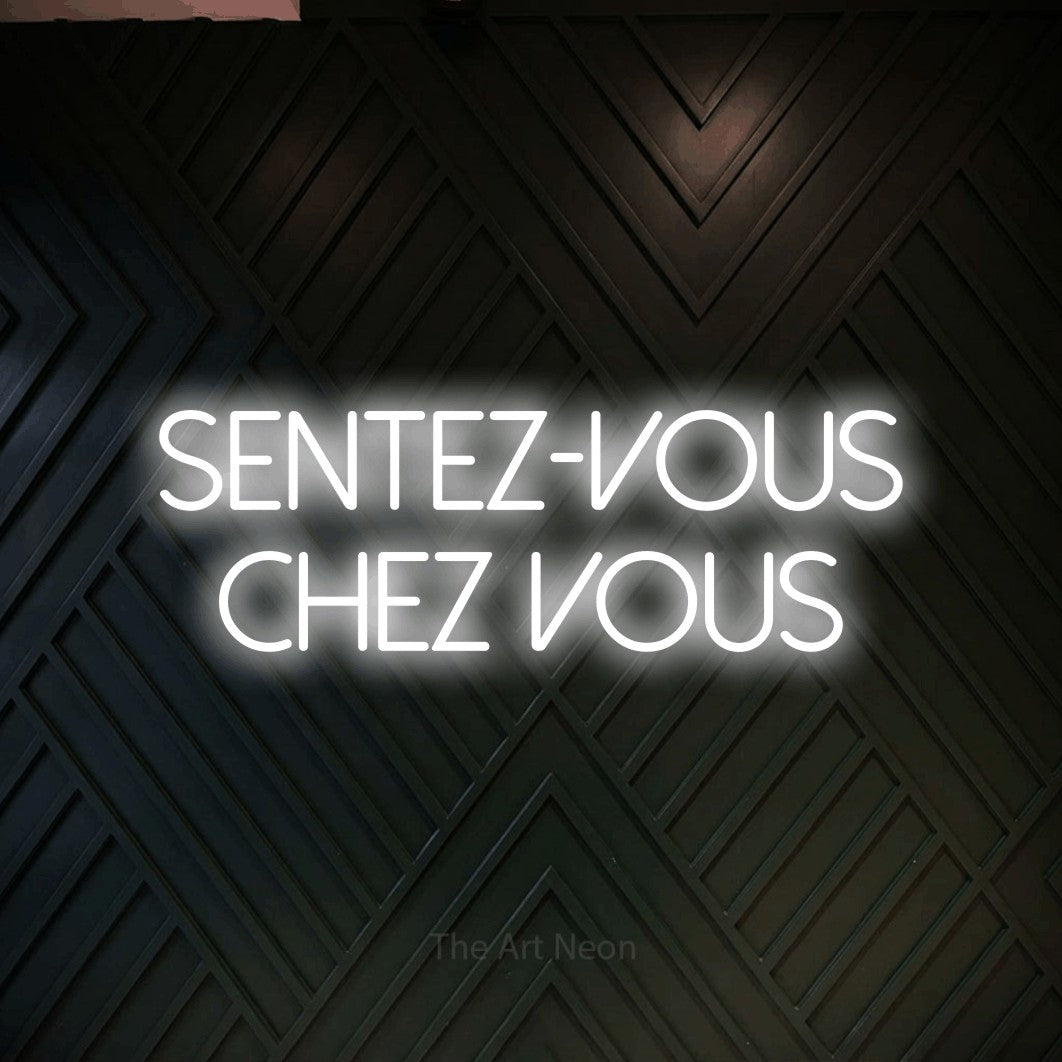 “Sentez-vous chez vous” LED neon sign in cool white, with thin sans-serif lettering across 2 lines, shown against a black chevron-patterned wood wall.