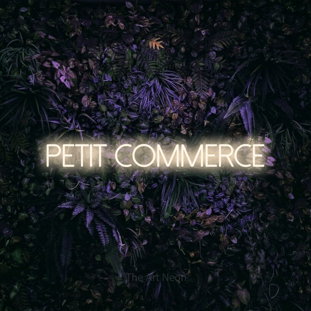 “Petit commerce” LED neon sign in warm white, with thin sans-serif lettering, shown against a dark wall with purple flowers.