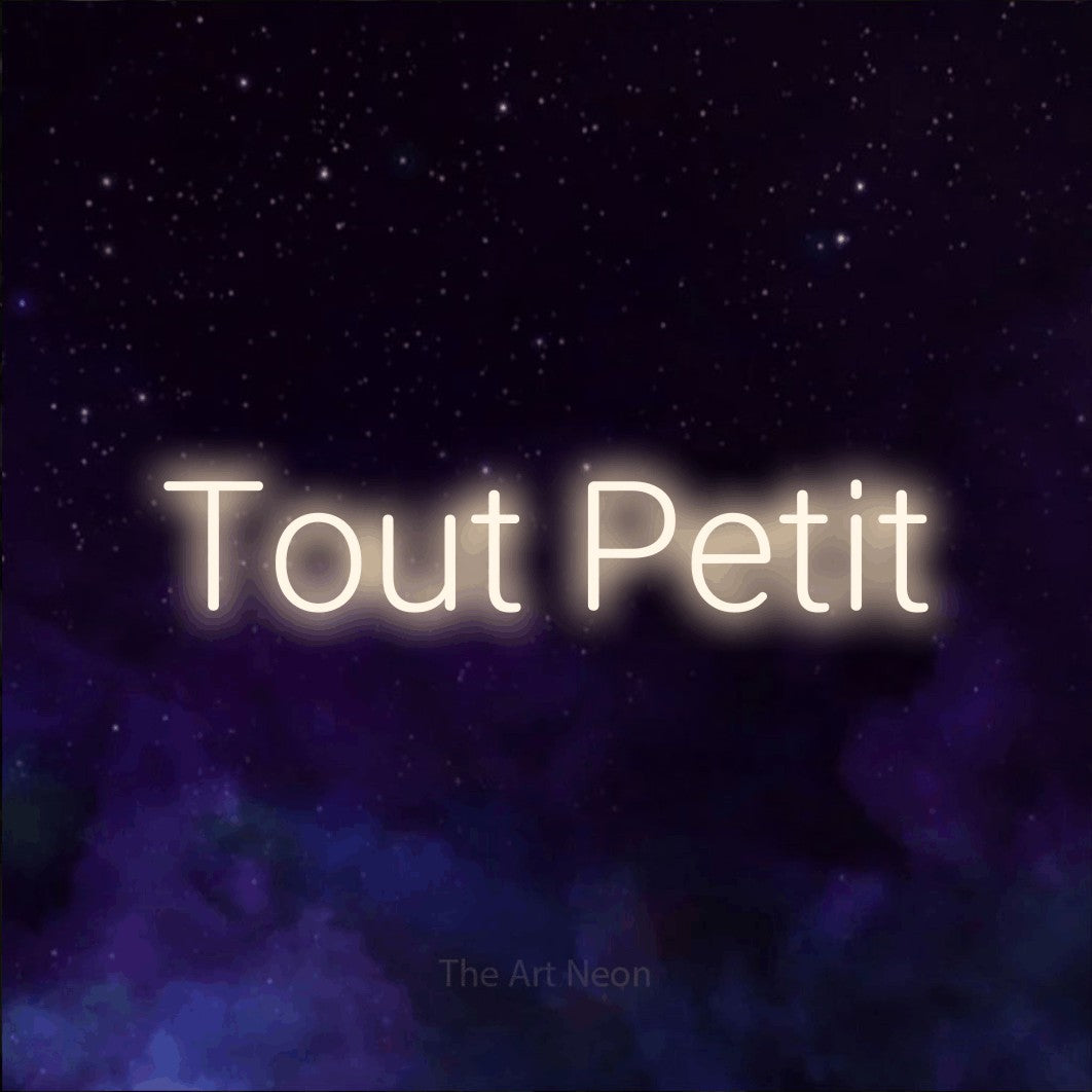 “Tout petit” LED neon sign in warm white, with rounded sans-serif lettering, shown against a purple galaxy backdrop.