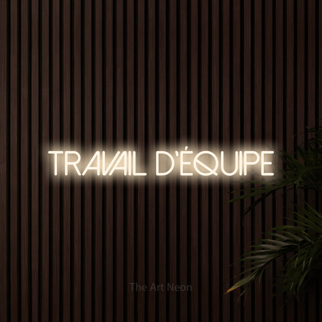 “Travail d'équipe” LED neon sign in warm white, with thin sans-serif lettering, shown against dark wooden wall slats.