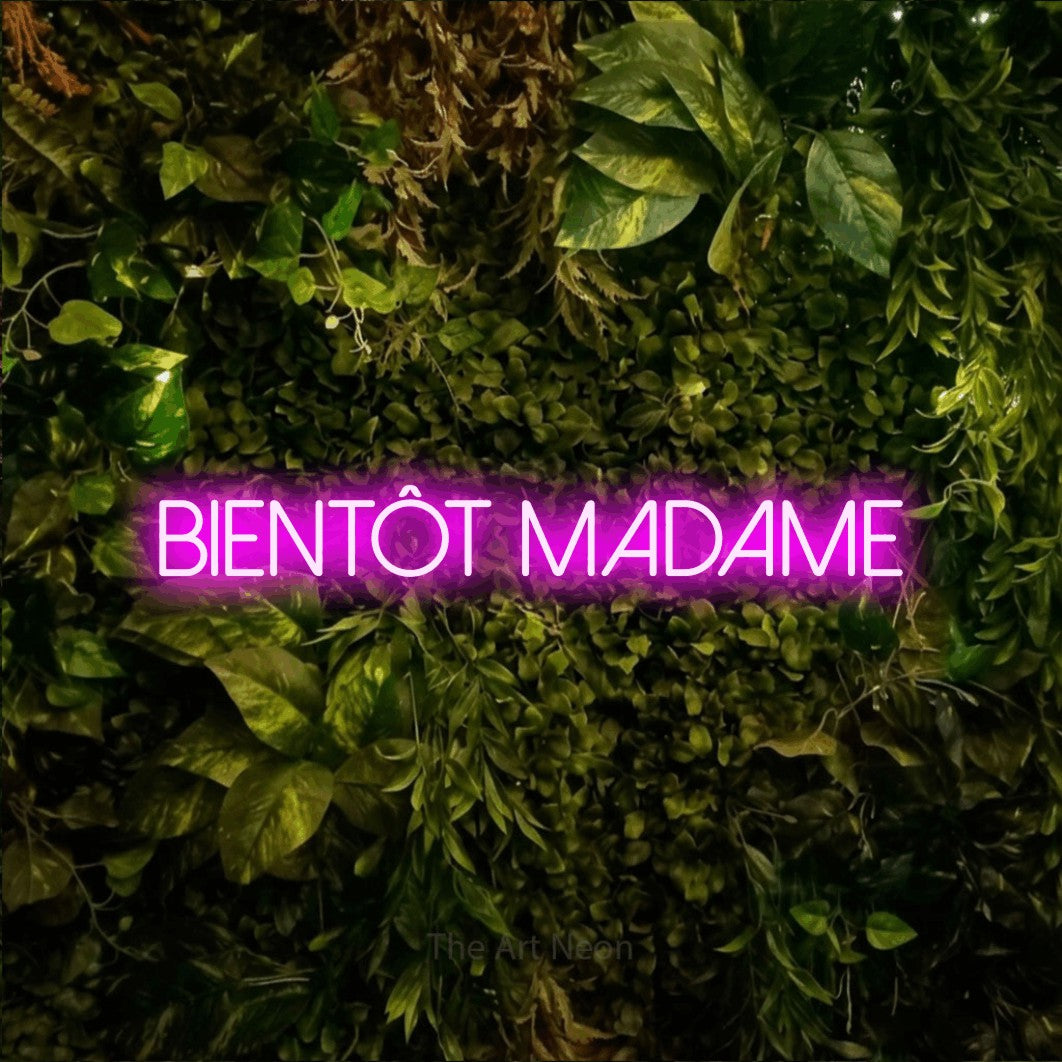 “Bientôt madame” LED neon sign in pink, with thin sans-serif lettering, shown against bright green and yellow foliage.