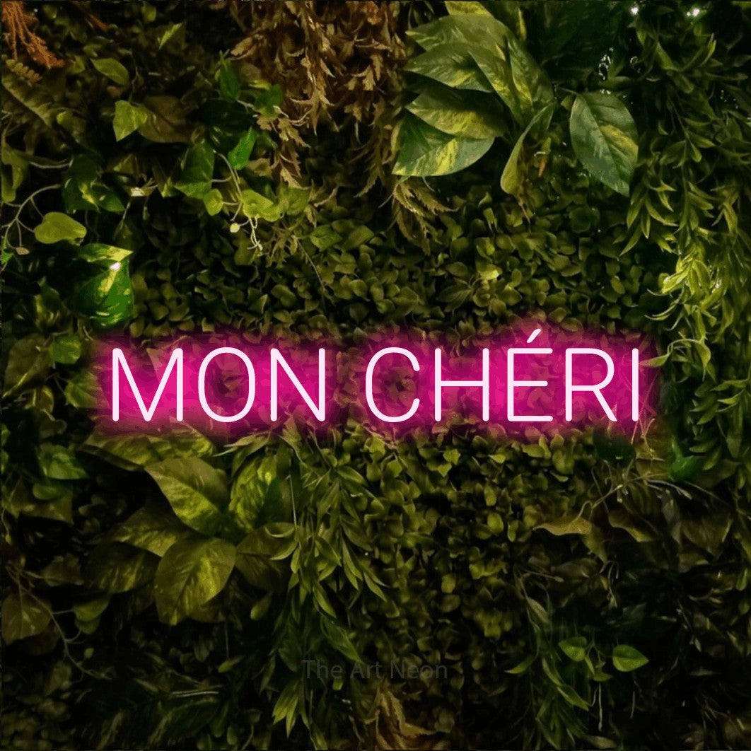 “Mon chéri” LED neon sign in hot pink, with rounded sans-serif lettering, shown against bright green and yellow foliage.