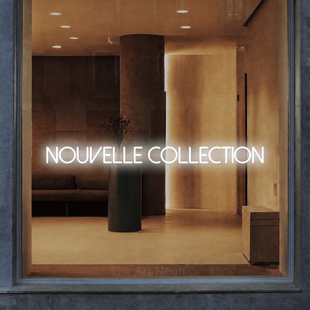 “Nouvelle collection” LED neon sign in cool white, with thin sans-serif lettering, shown against a warm-toned interior.