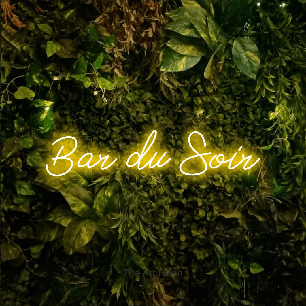 Décoration murale en néon LED « Bar du soir » en jaune, en lettrage cursif, installée sur un feuillage vert et jaune vif, pour un bar ou un pub.
