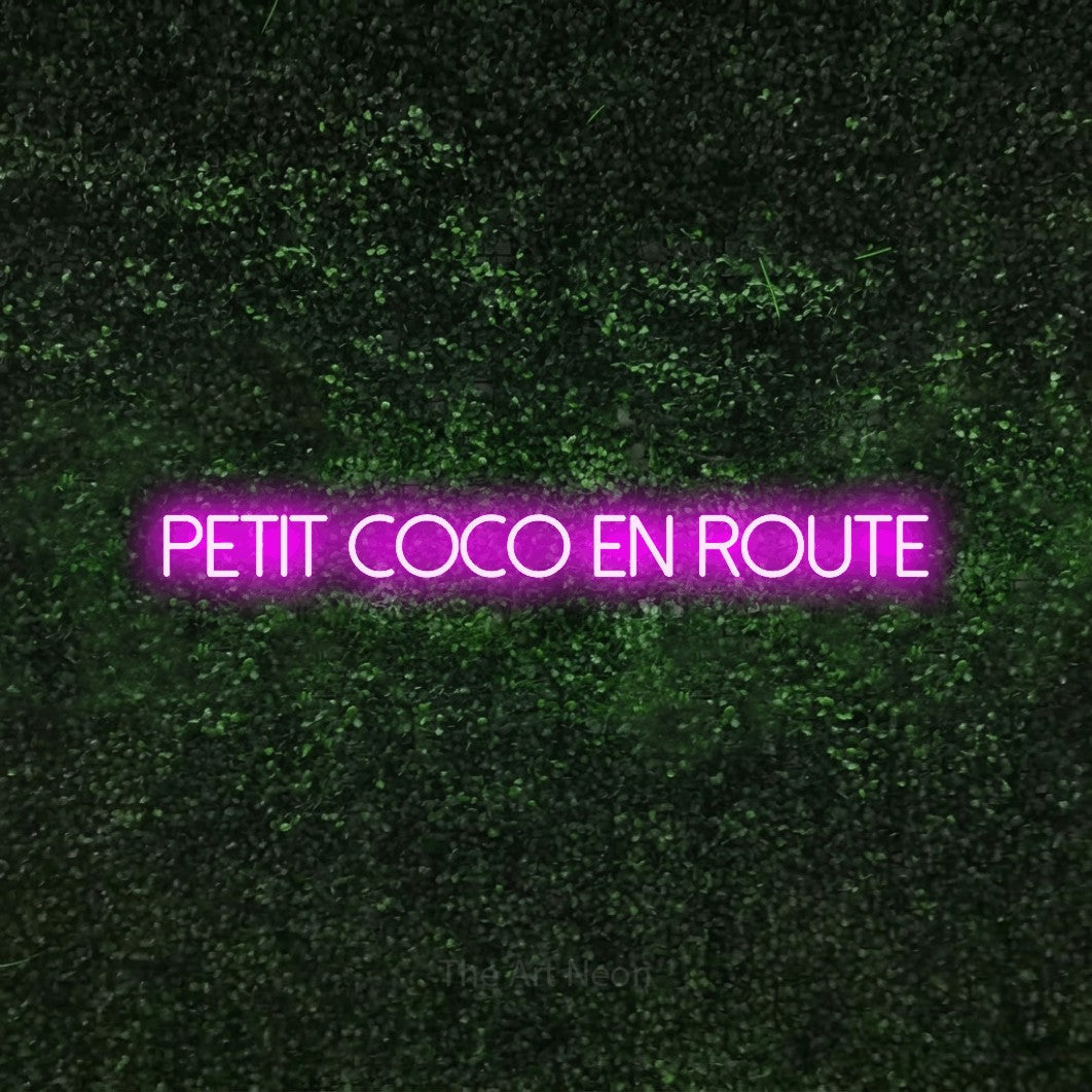 Décoration murale en néon LED « Petit coco en route » en rose, en lettres fines sans empattement, installée sur une haie verte texturée, pour une fête prénatale.
