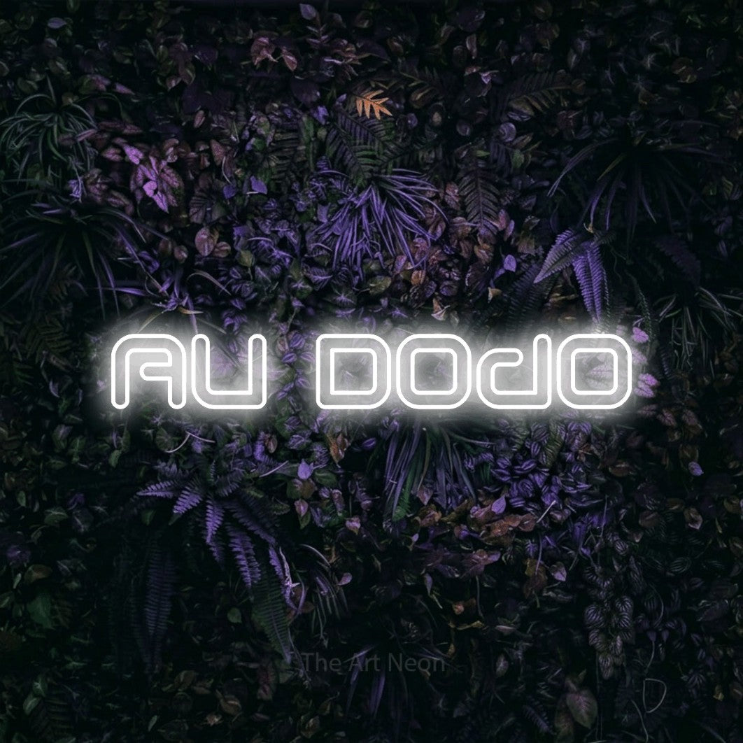“Au dodo” LED neon sign in cool white, with futuristic double-line lettering, shown against a dark wall with purple flowers.