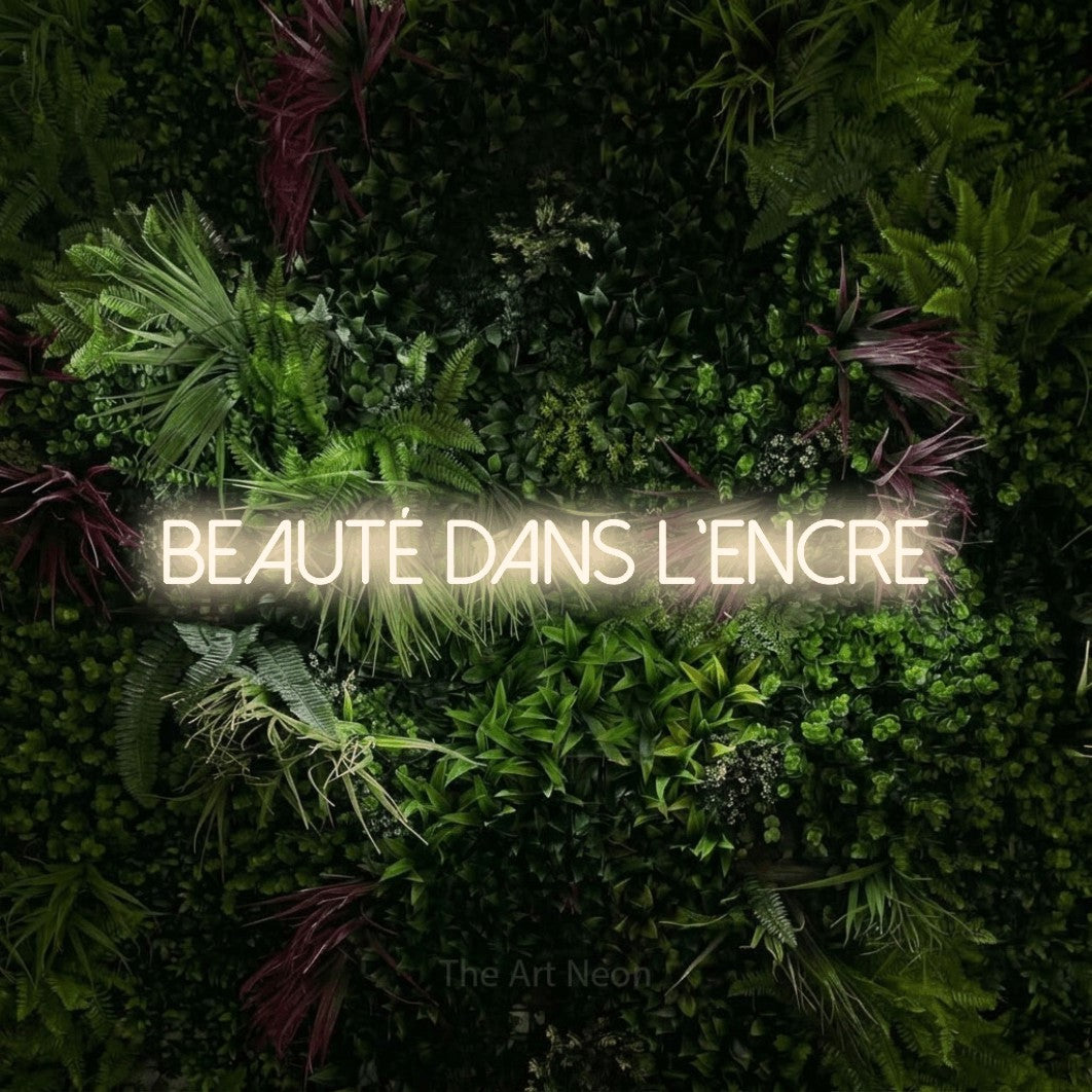 “Beauté dans l'encre” LED neon sign in warm white, with thin sans-serif lettering, shown against a plant wall with pink flowers.
