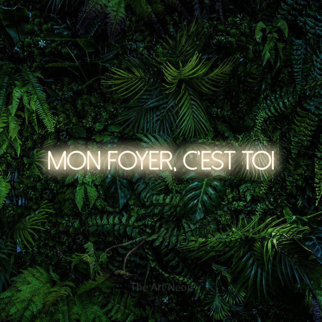 “Mon foyer, c'est toi” LED neon sign in warm white, with thin sans-serif lettering, shown against dense tropical foliage.