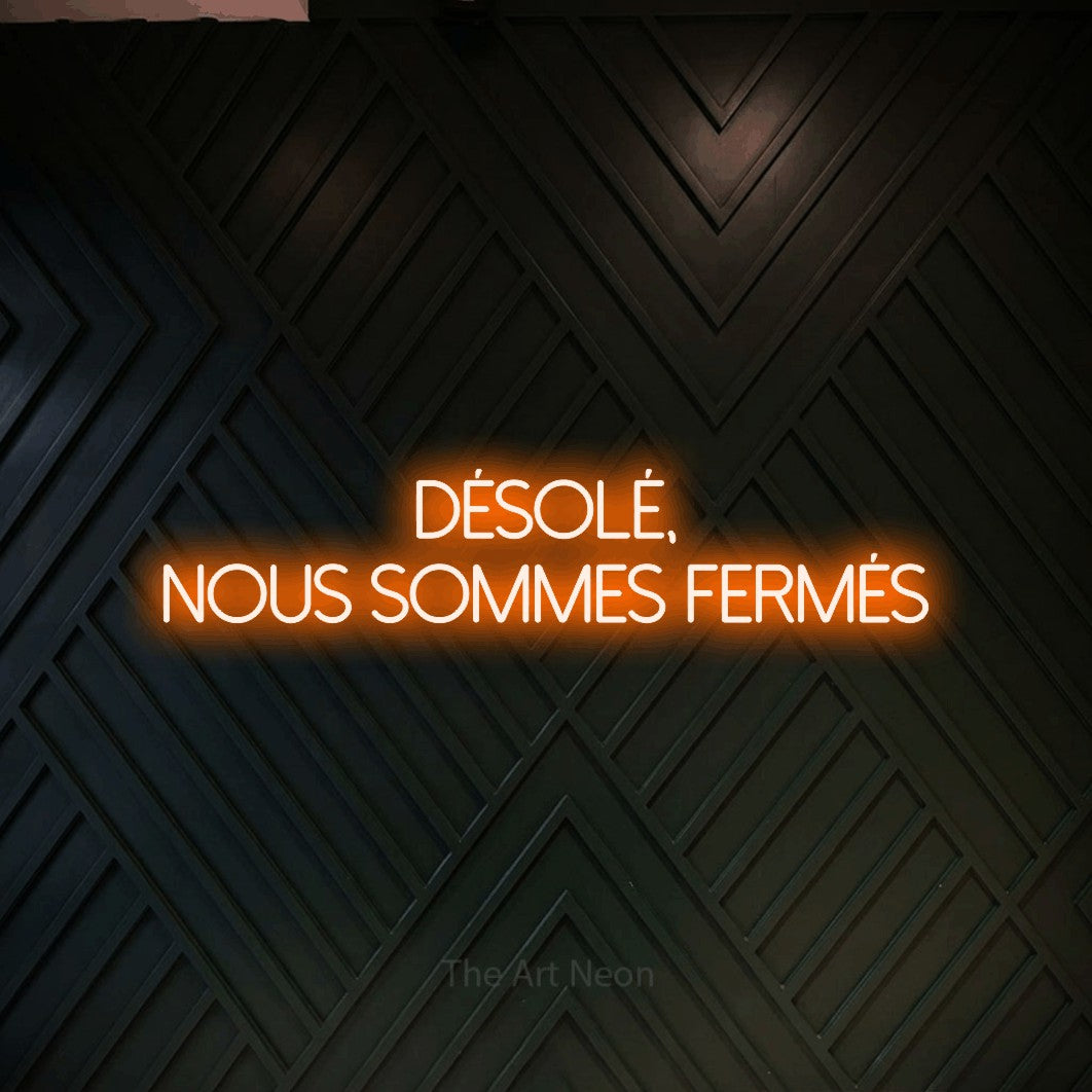 “Désolé, nous sommes fermés” LED neon sign in orange, with thin sans-serif lettering across 2 lines, shown against a black chevron-patterned wood wall.