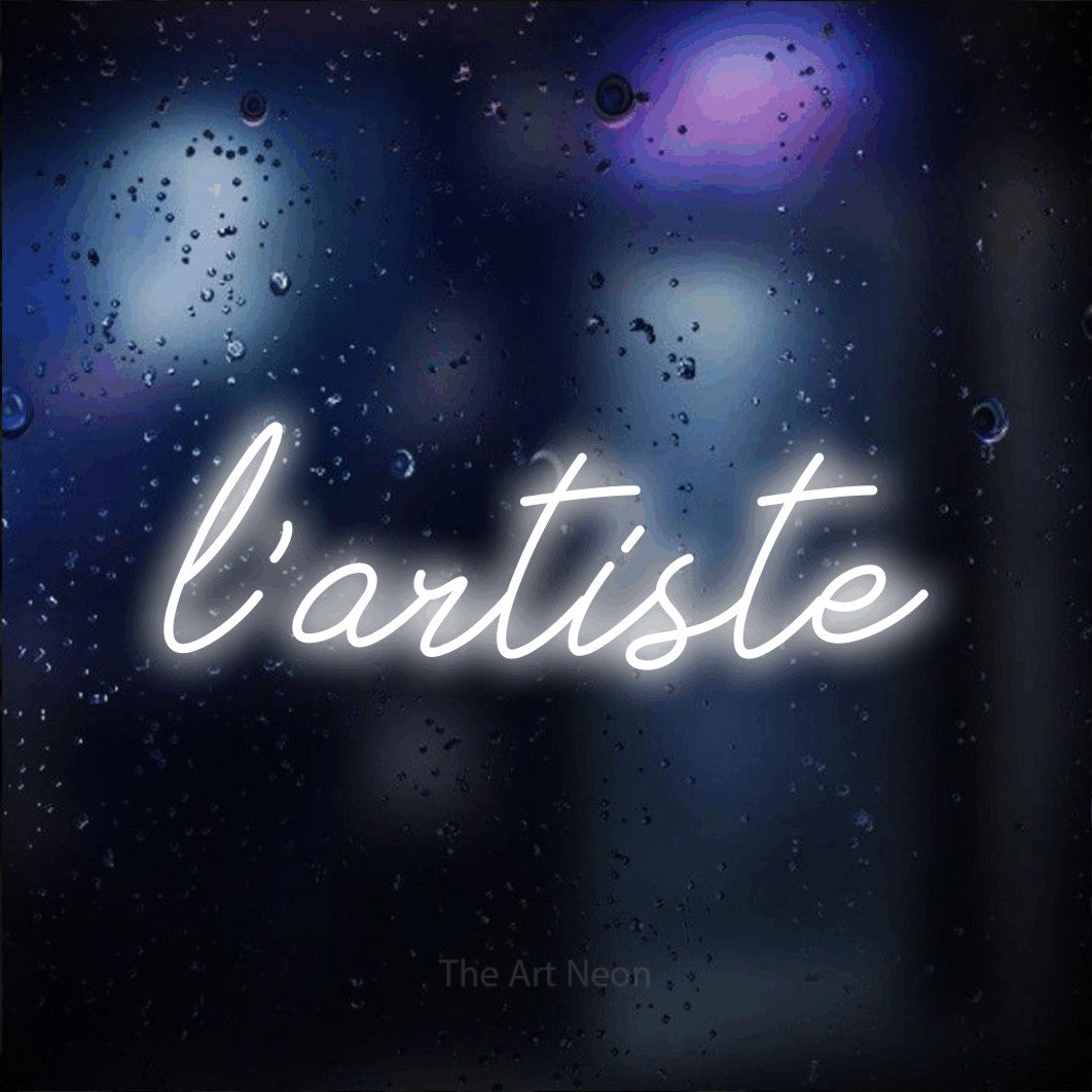 “L'artiste” LED neon sign in cool white, with cursive lettering, shown against glass with blue and purple reflections.