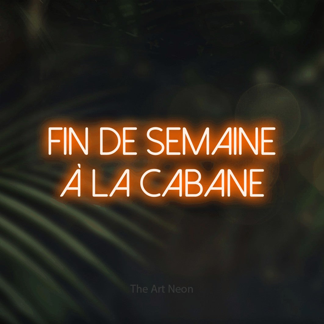 “Fin de semaine à la cabane” LED neon sign in orange, with thin sans-serif lettering across 2 lines, shown against a blurred palm and bokeh-light backdrop.