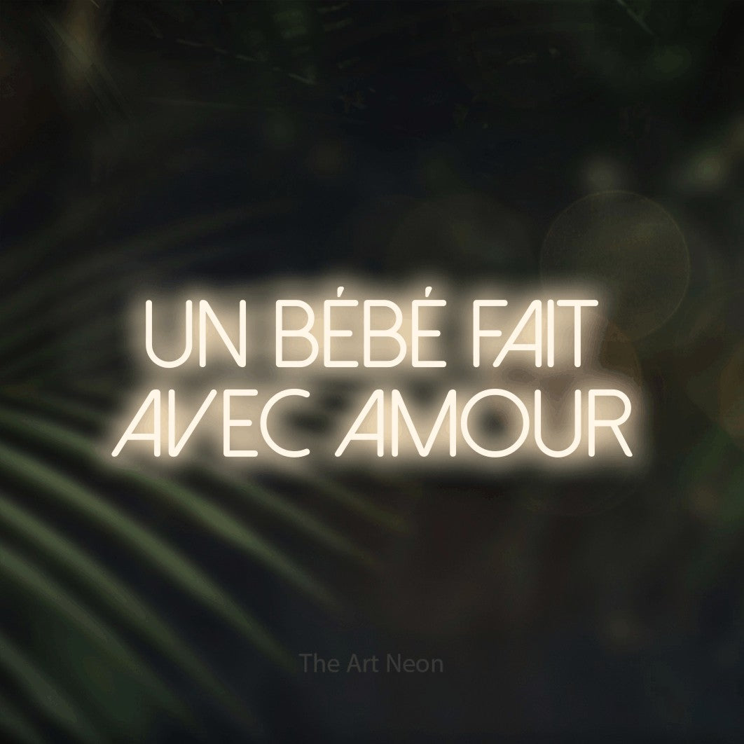“Un bébé fait avec amour” LED neon sign in warm white, with thin sans-serif lettering across 2 lines, shown against a blurred palm and bokeh-light backdrop.