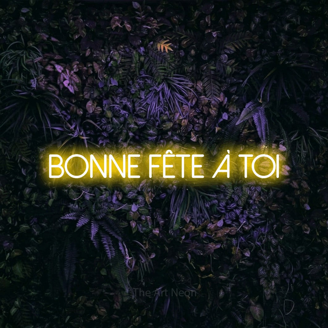“Bonne fête à toi” LED neon sign in yellow, with thin sans-serif lettering, shown against a dark wall with purple flowers.