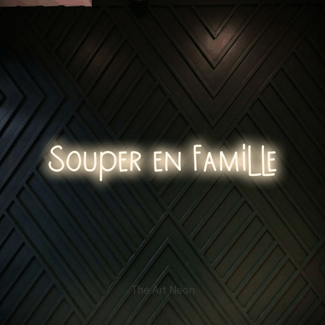 “Souper en famille” LED neon sign in warm white, with narrow hand-drawn lettering, shown against a black chevron-patterned wood wall.