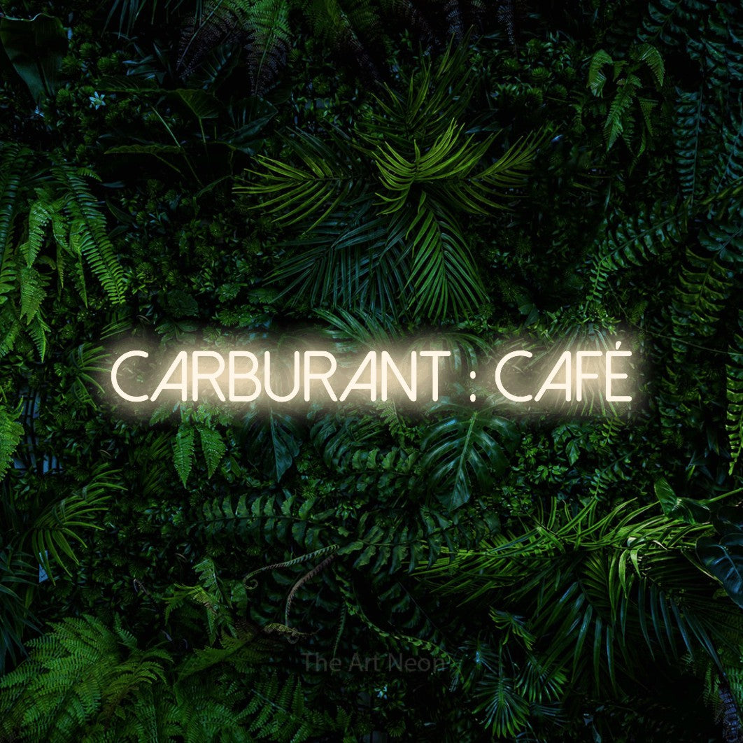 “Carburant : café” LED neon sign in warm white, with thin sans-serif lettering, shown against dense tropical foliage.