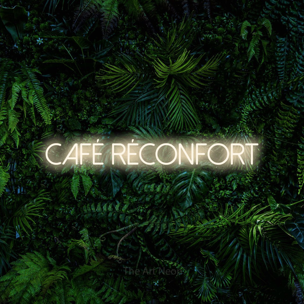 “Café réconfort” LED neon sign in warm white, with thin sans-serif lettering, shown against dense tropical foliage.