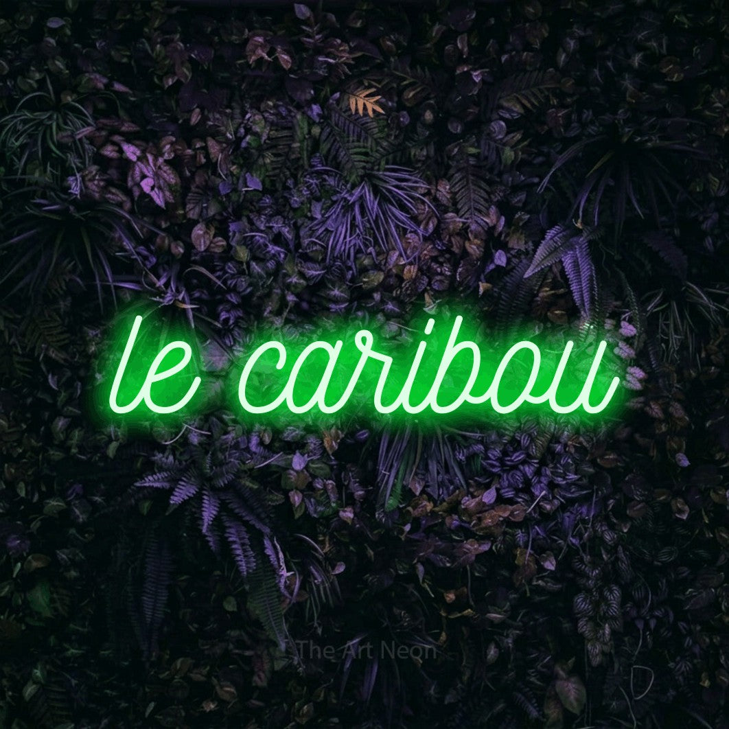“Le caribou” LED neon sign in green, with ornate cursive lettering, shown against a dark wall with purple flowers.