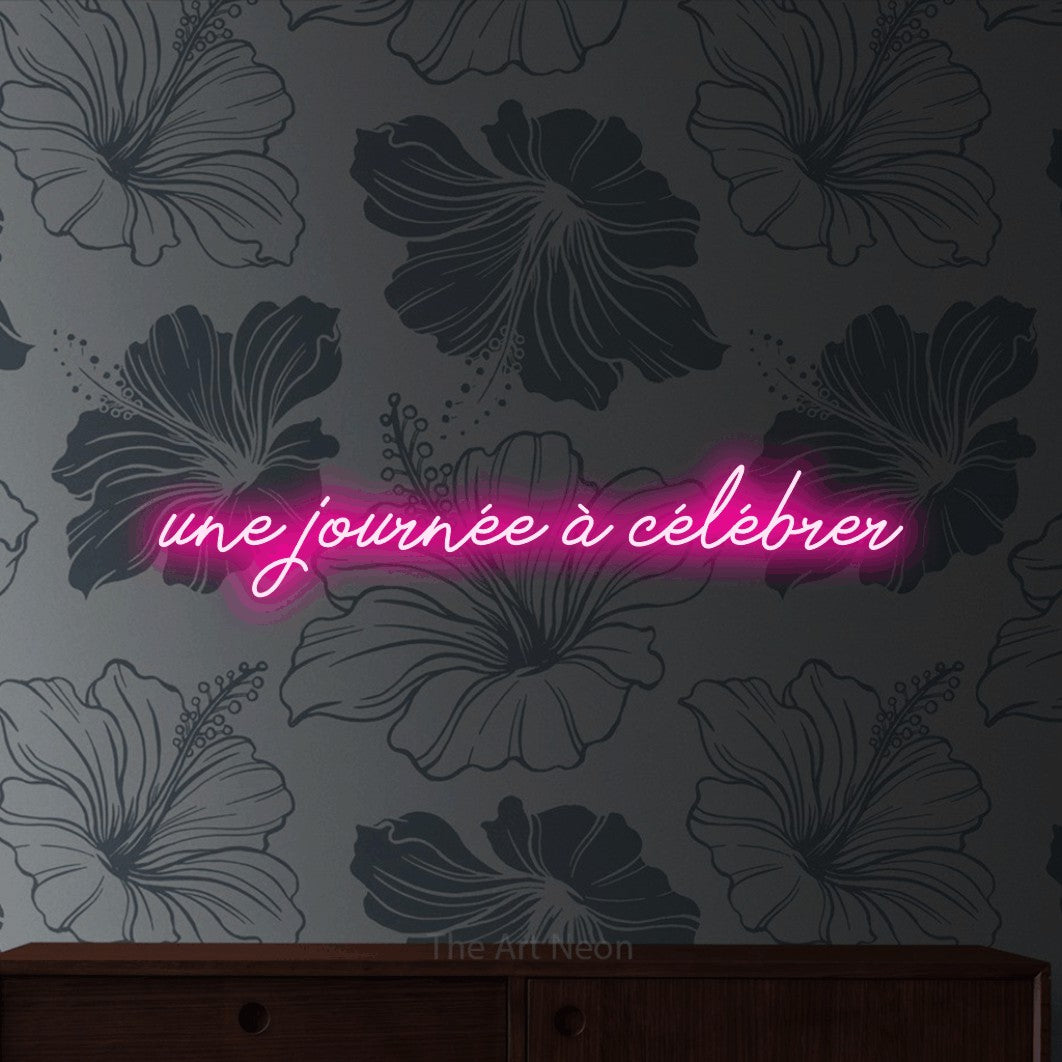 “Une journée à célébrer” LED neon sign in hot pink, with cursive lettering, shown against a grey floral-patterned wall.