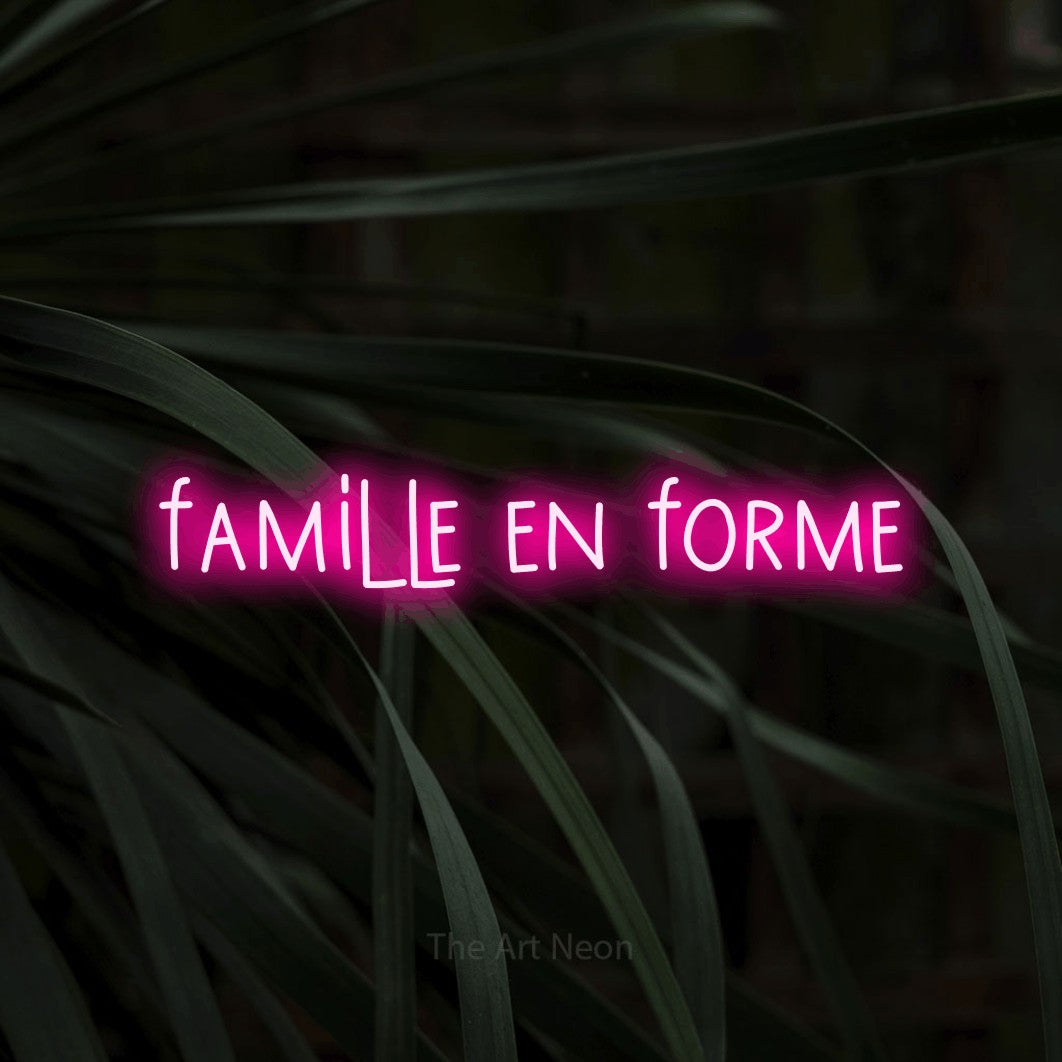 Aperçu d’une enseigne néon LED « Famille en forme » en rose vif, en lettres étroites dessinées à la main, photographiée sur un feuillage sombre en gros plan, pour un espace d’entraînement.