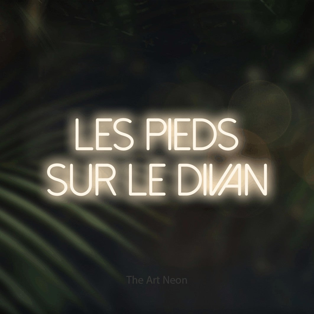 Composition en néon LED « Les pieds sur le divan » sur deux lignes en blanc chaud, en lettres fines sans empattement, mise en valeur sur un fond flou de palmiers et de lumières bokeh, pour un salon.