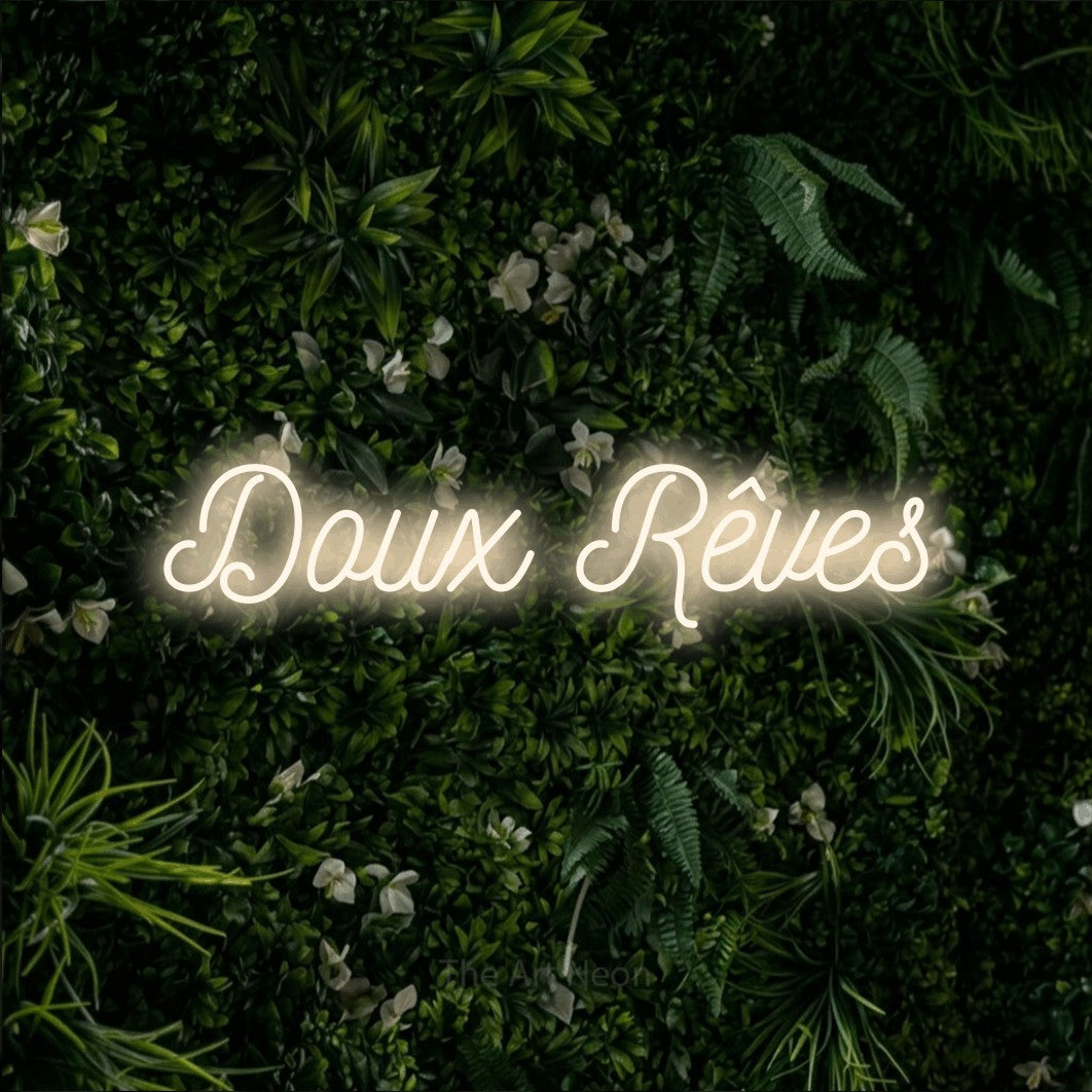“Doux rêves” LED neon sign in warm white, with ornate cursive lettering, shown against a dense green plant wall.