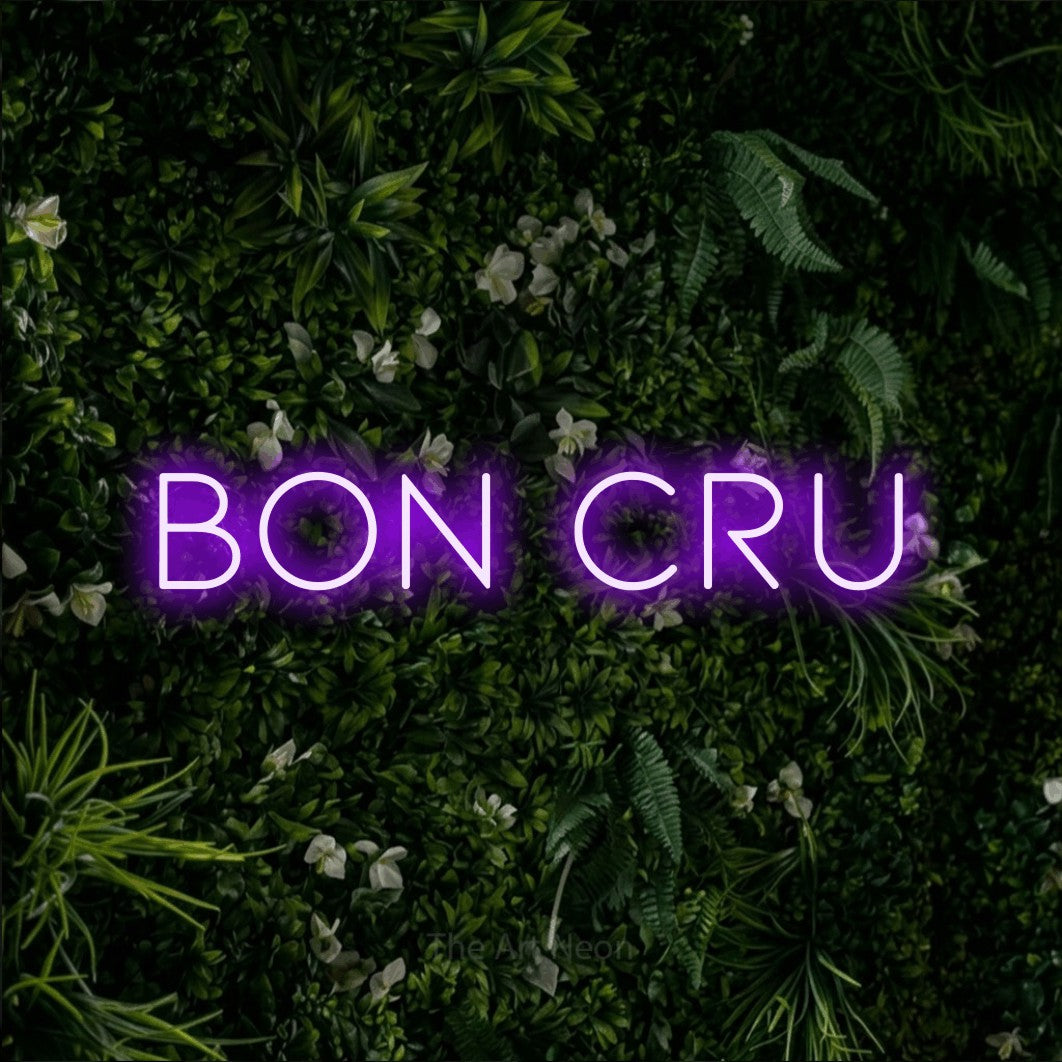 “Bon cru” LED neon sign in purple, with thin, widely spaced lettering, shown against a dense green plant wall.
