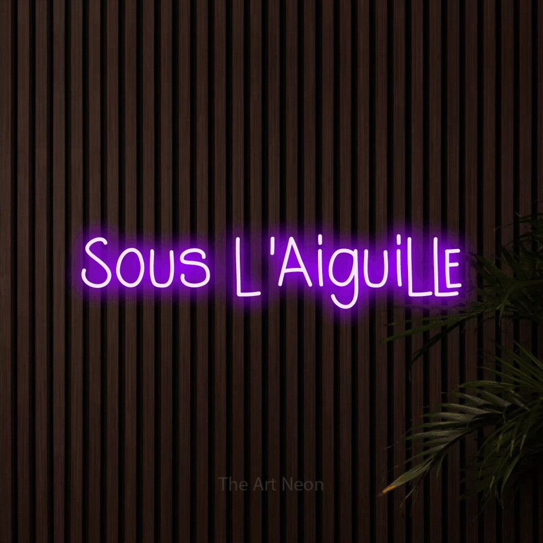 “Sous l'aiguille” LED neon sign in purple, with narrow hand-drawn lettering, shown against dark wooden wall slats.