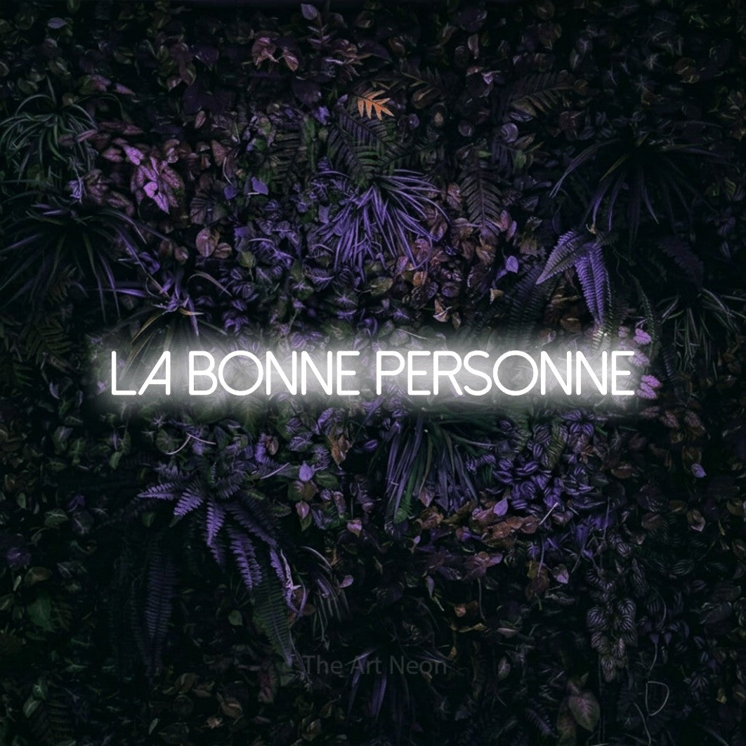 “La bonne personne” LED neon sign in cool white, with thin sans-serif lettering, shown against a dark wall with purple flowers.
