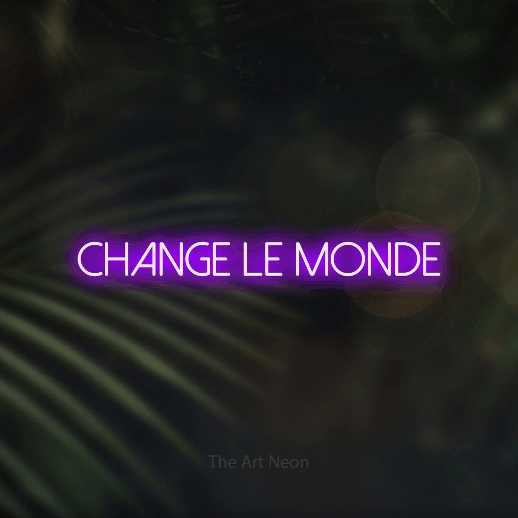 “Change le monde” LED neon sign in purple, with thin sans-serif lettering, shown against a blurred palm and bokeh-light backdrop.