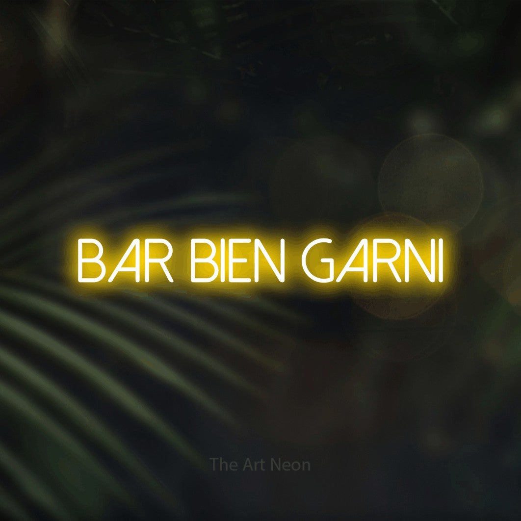 “Bar bien garni” LED neon sign in yellow, with thin sans-serif lettering, shown against a blurred palm and bokeh-light backdrop.