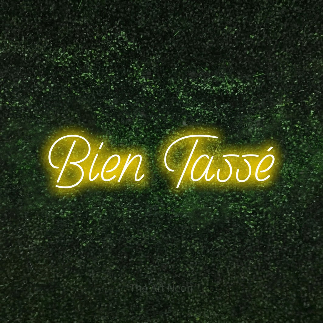 “Bien tassé” LED neon sign in yellow, with slanted handwritten lettering, shown against a textured green hedge.