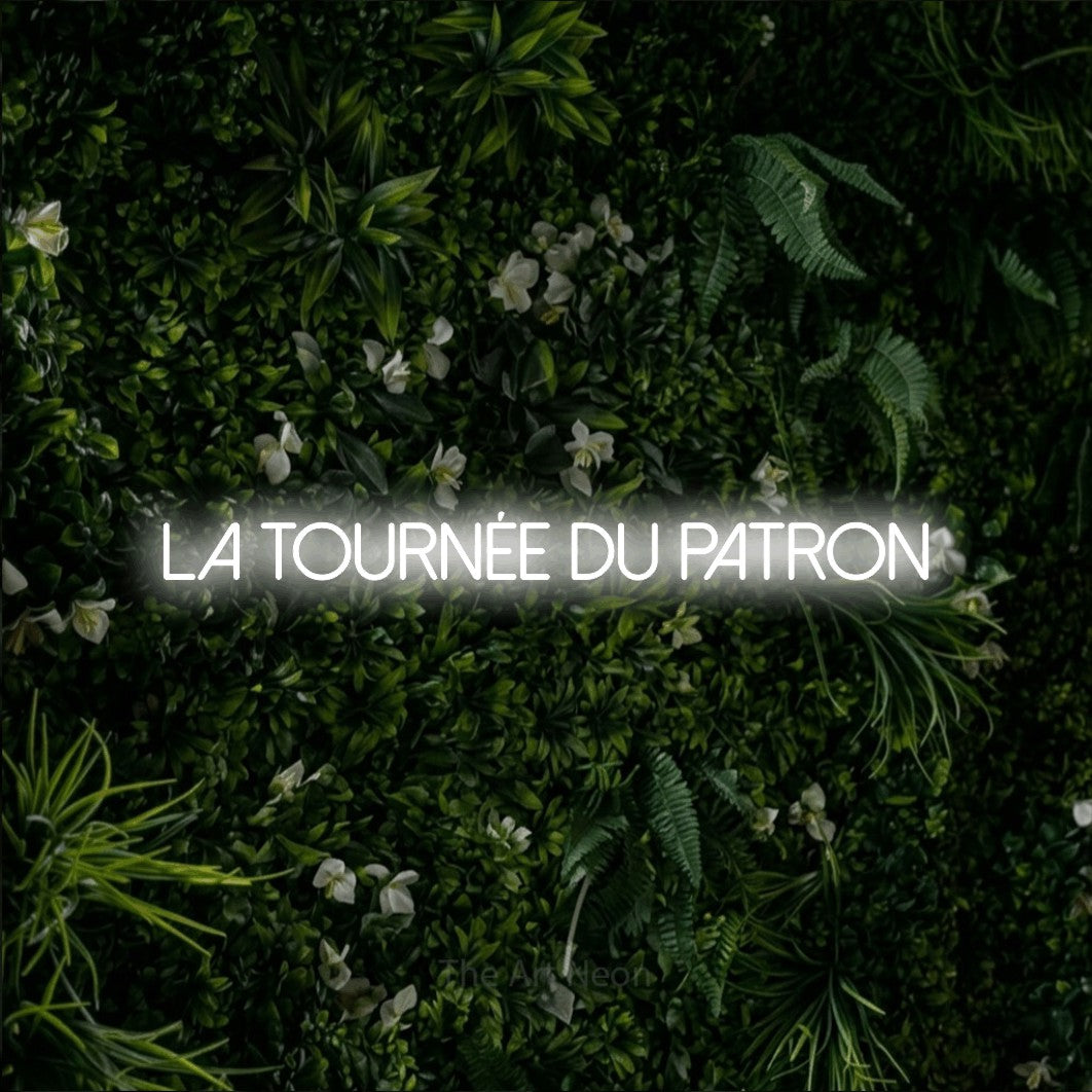 Lettrage en néon LED « La tournée du patron » en blanc froid, en lettres fines sans empattement, présenté sur un mur végétal vert et dense, pour un bar ou un pub.